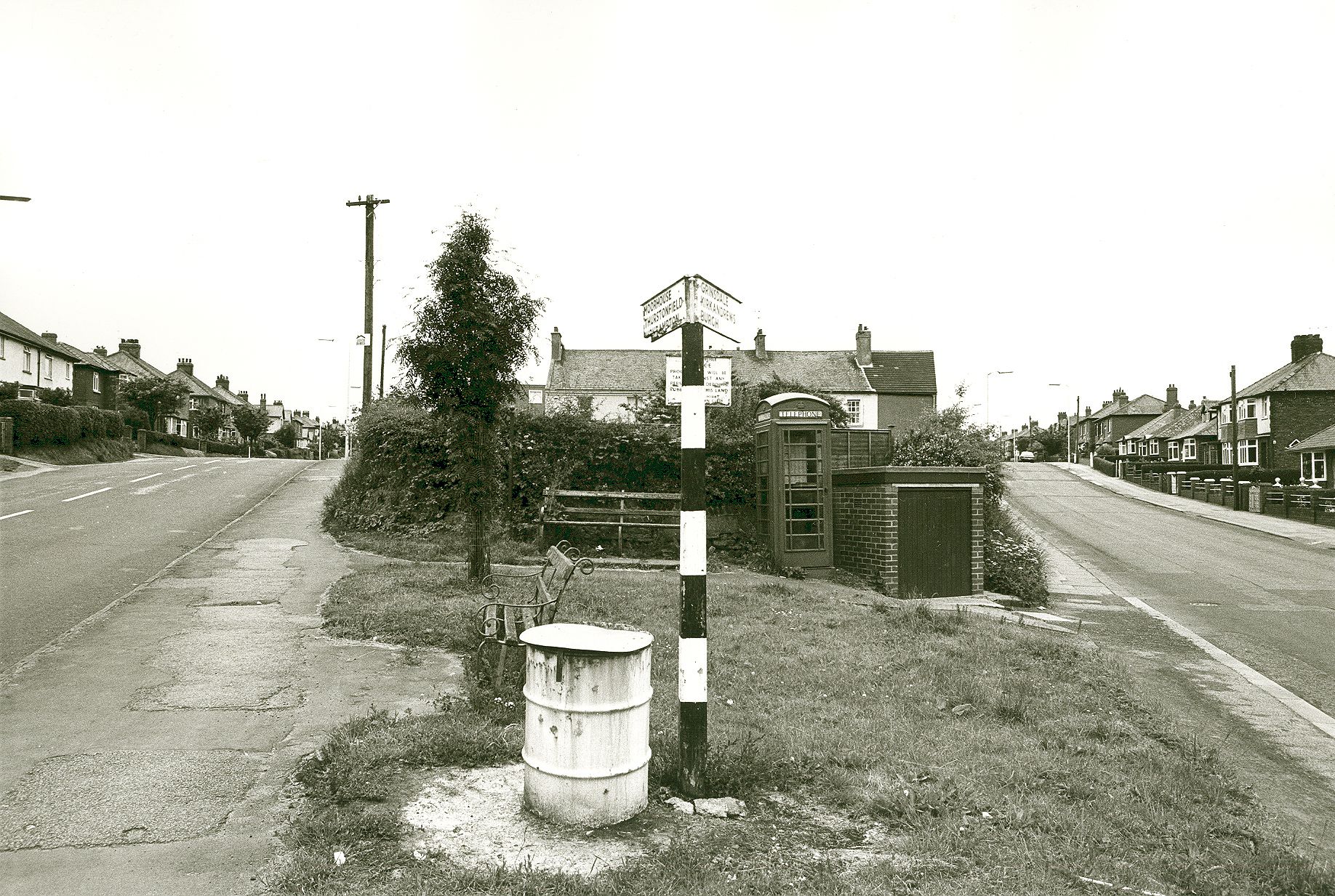 A black-and-white photograph of a road fork with a signpost pointing left to Moorhouse Road and right to Burgh Road, and an old telephone box beside it
