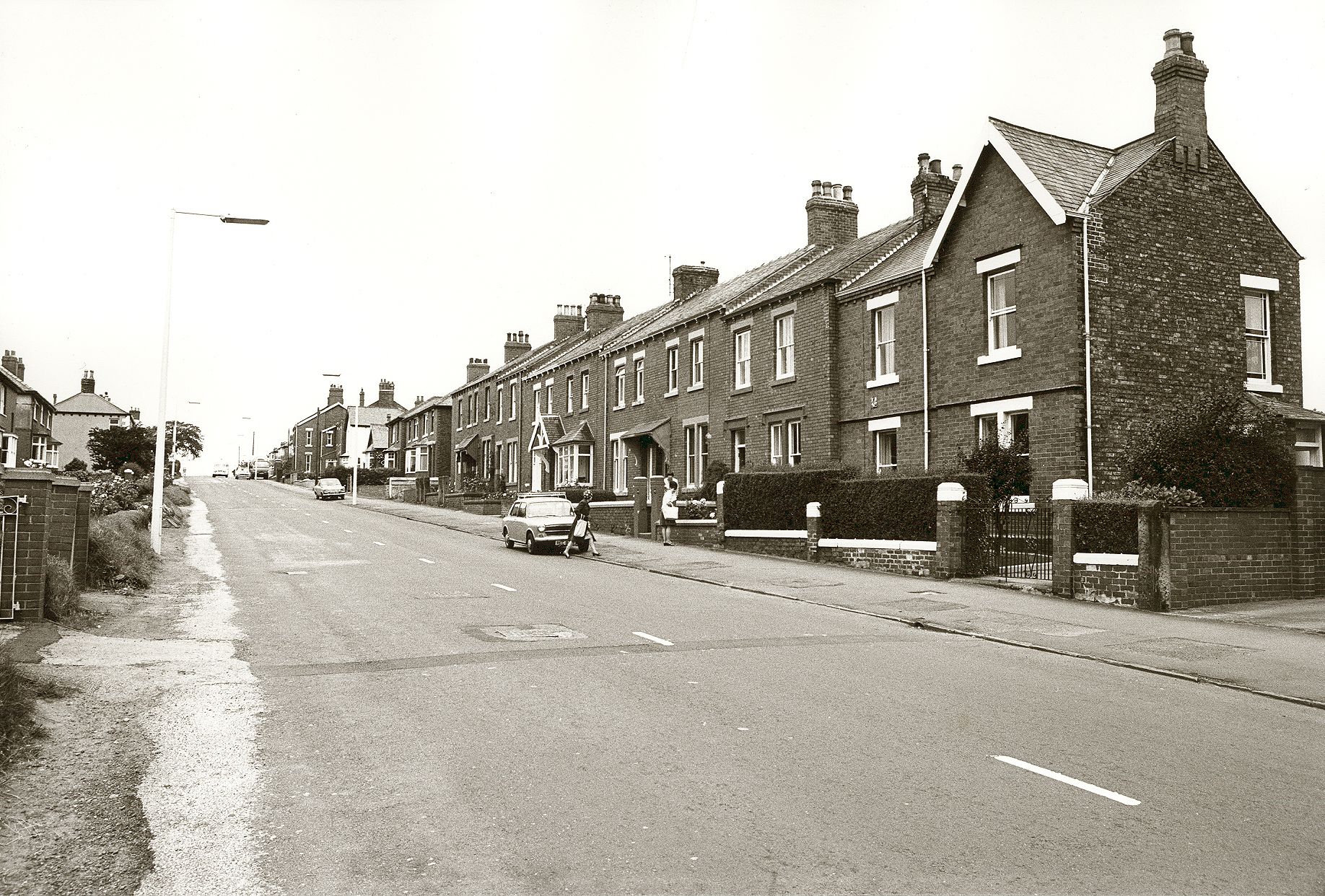 A black-and-white photograph of a long terrace of brick houses on Moorhouse Road, with a family getting out of a car in the foreground