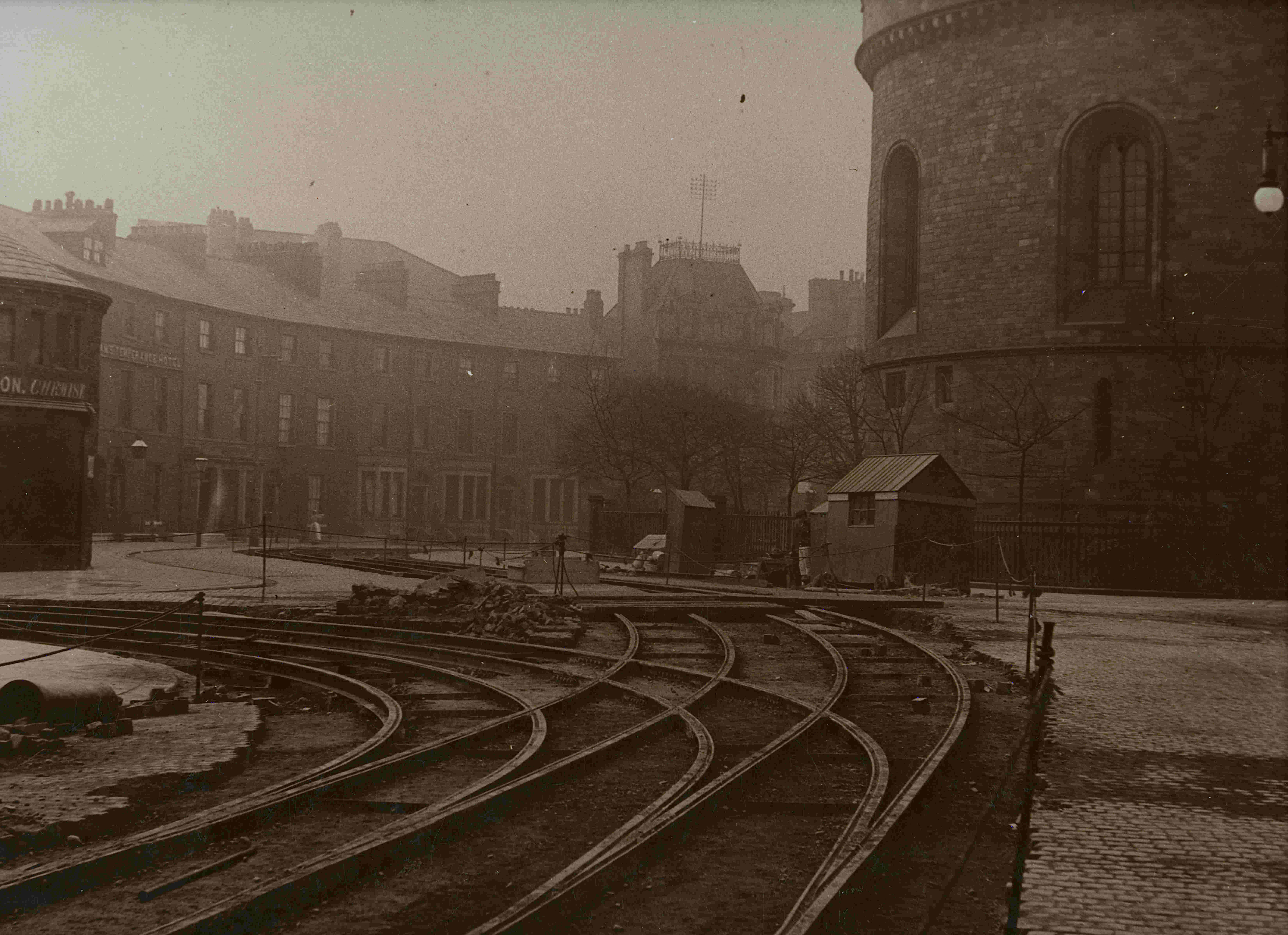 A sepia atmospheric photograph of curved tram tracks running through the centre of Carlisle, with a stone church tower to the right