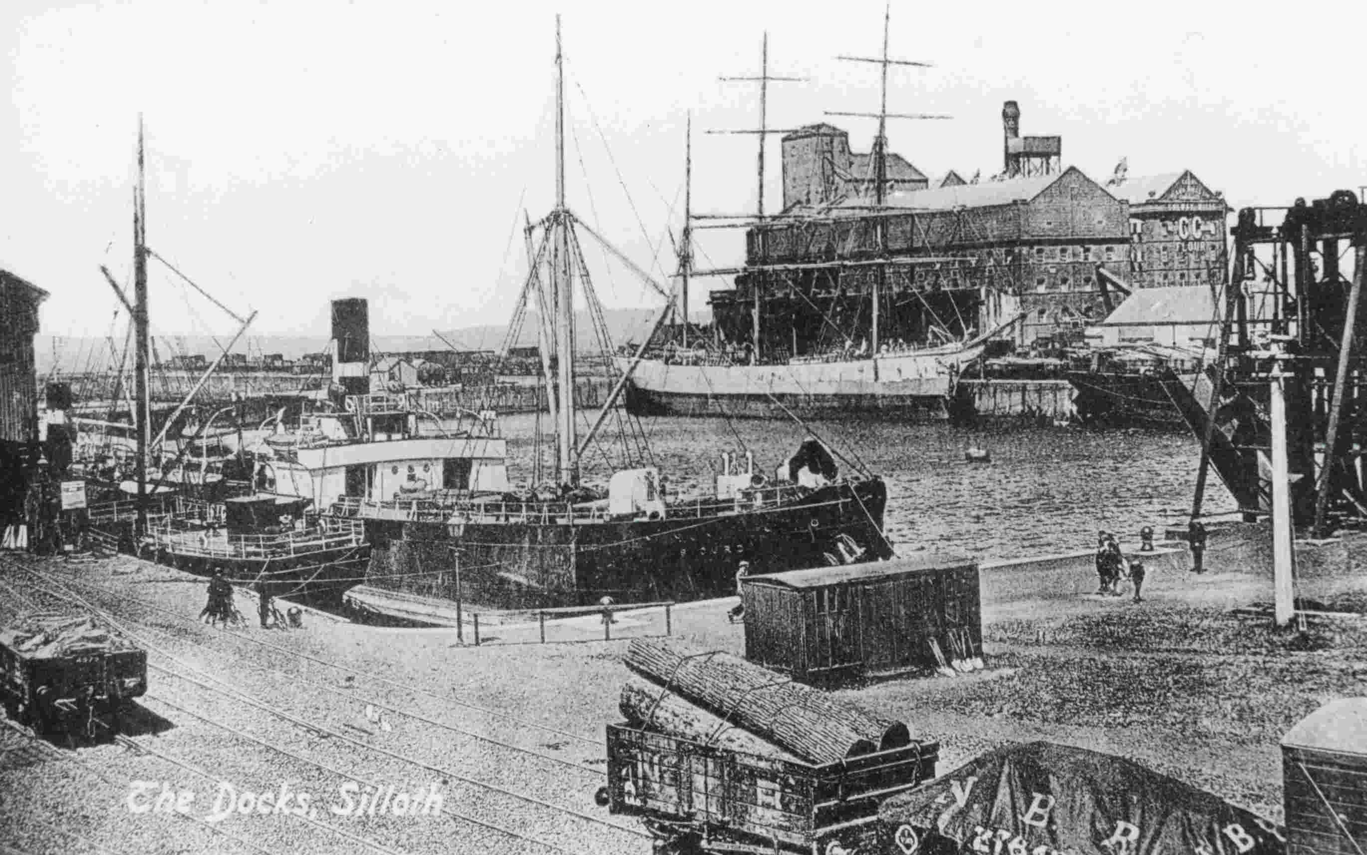 A black-and-white postcard of The Docks at Silloth, with several vessels moored alongside freight wagons on the quayside