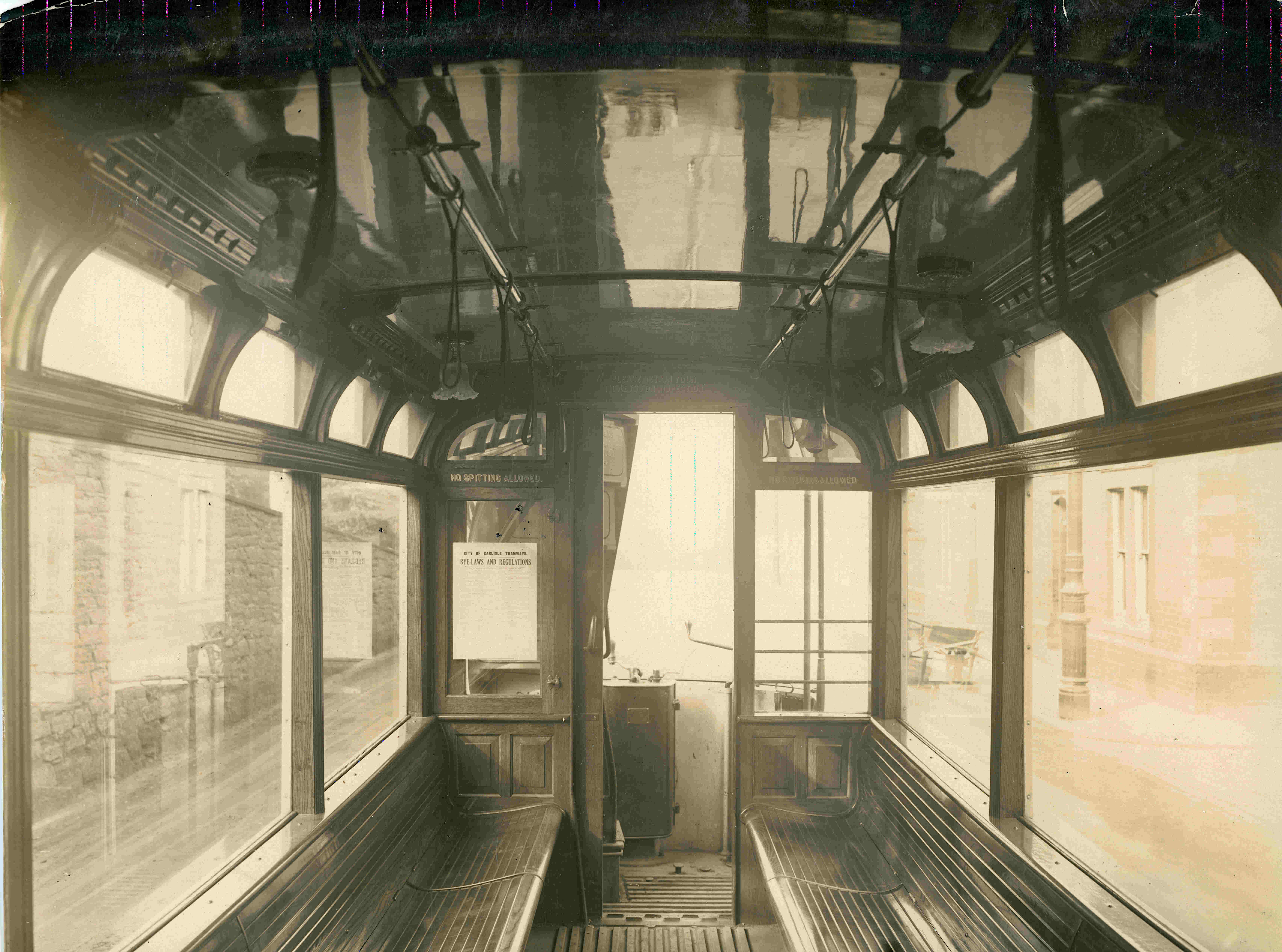 A sepia photograph of the interior of a City of Carlisle Tramways tram car, with wooden benches, arched windows and lamps on the ceiling