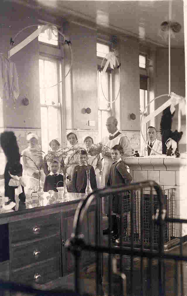 A sepia photograph of a hospital ward with doctors, nurses and children gathered, medical equipment visible and an iron cot in the foreground