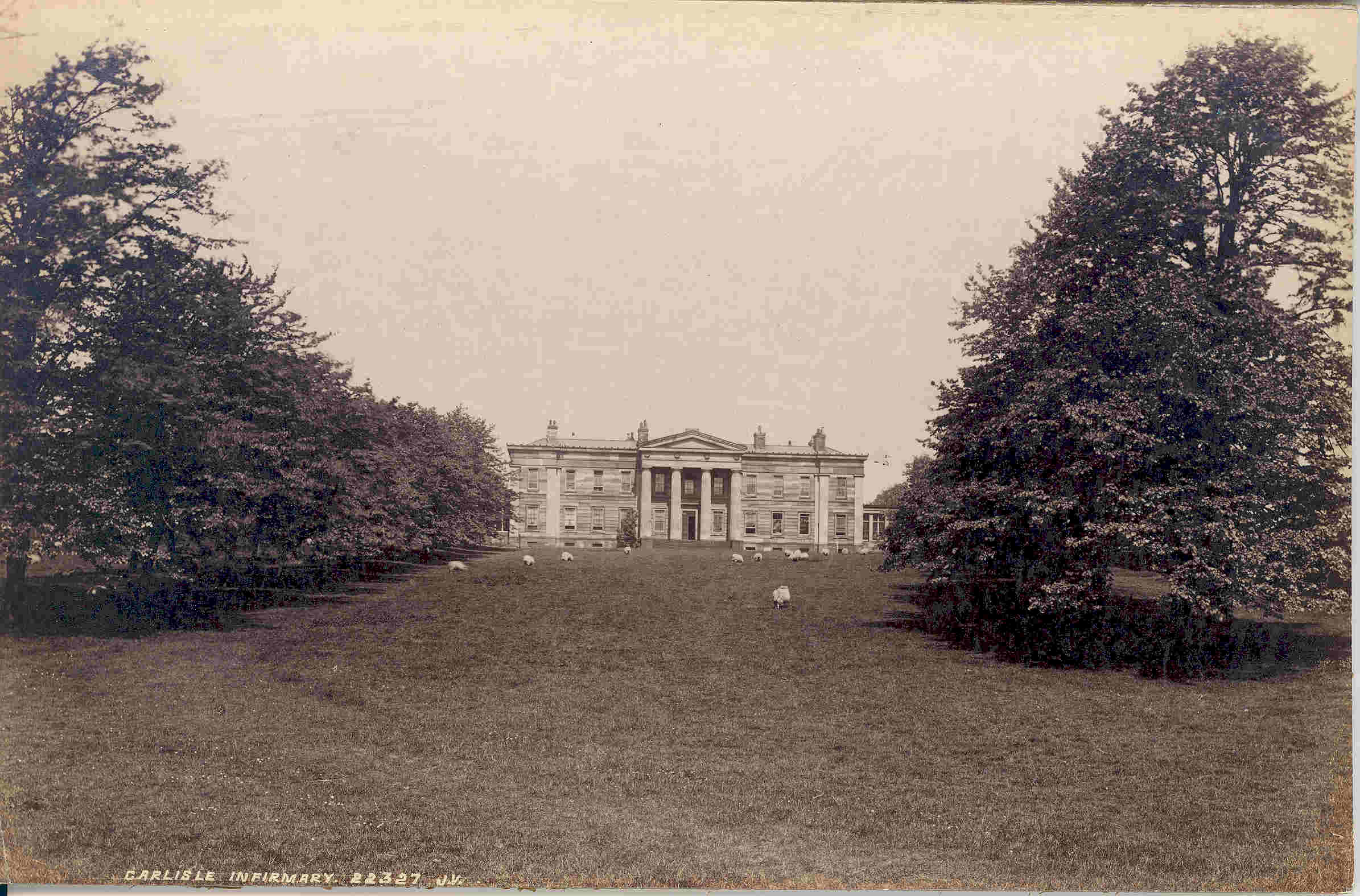 A sepia postcard of the original Cumberland Infirmary, a Georgian mansion seen across a wide lawn with sheep grazing in front, labelled Carlisle Infirmary