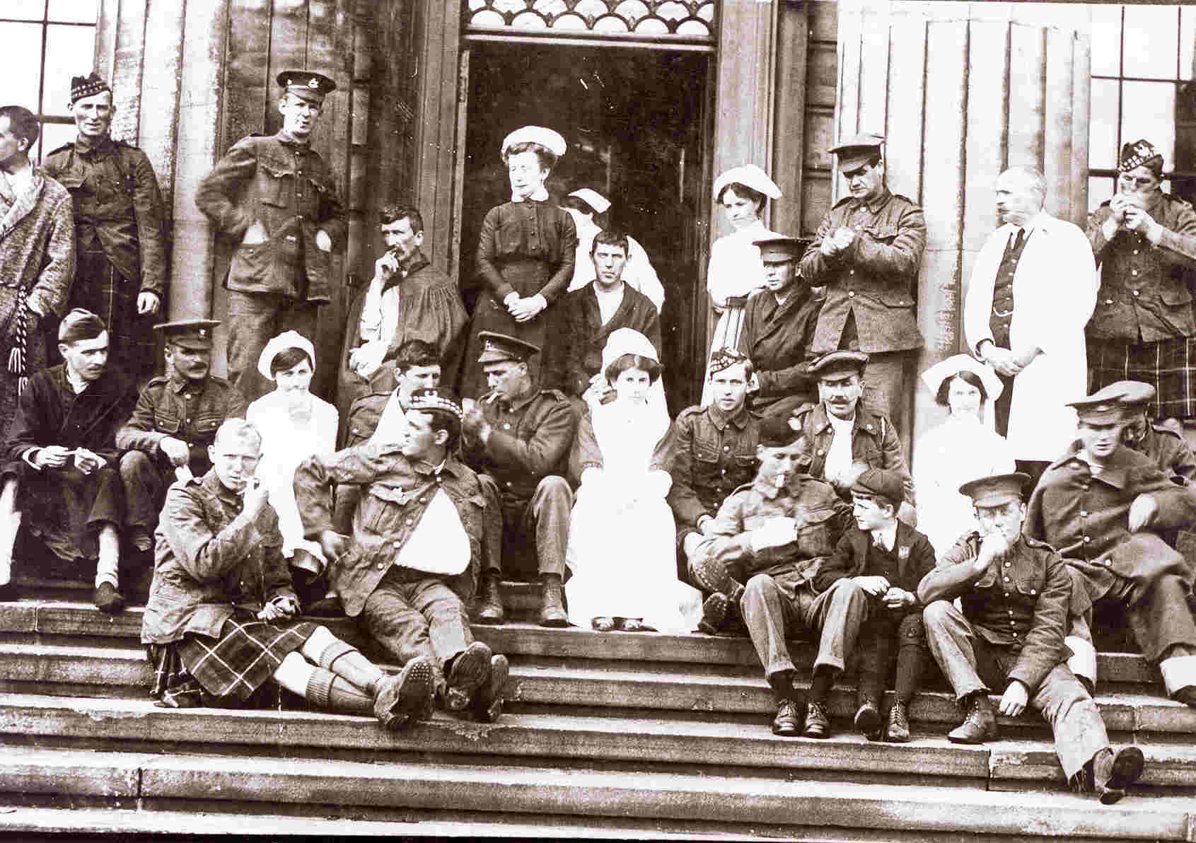 A sepia photograph of WW1 soldiers in uniform sitting on the infirmary steps, with nurses in white aprons among them