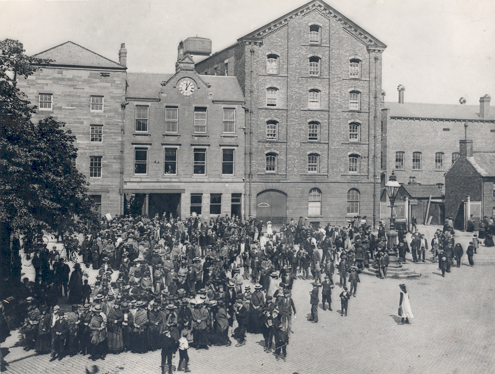 A Victorian photograph taken from above of hundreds of workers gathered in the factory yard in front of large brick buildings with a clock tower