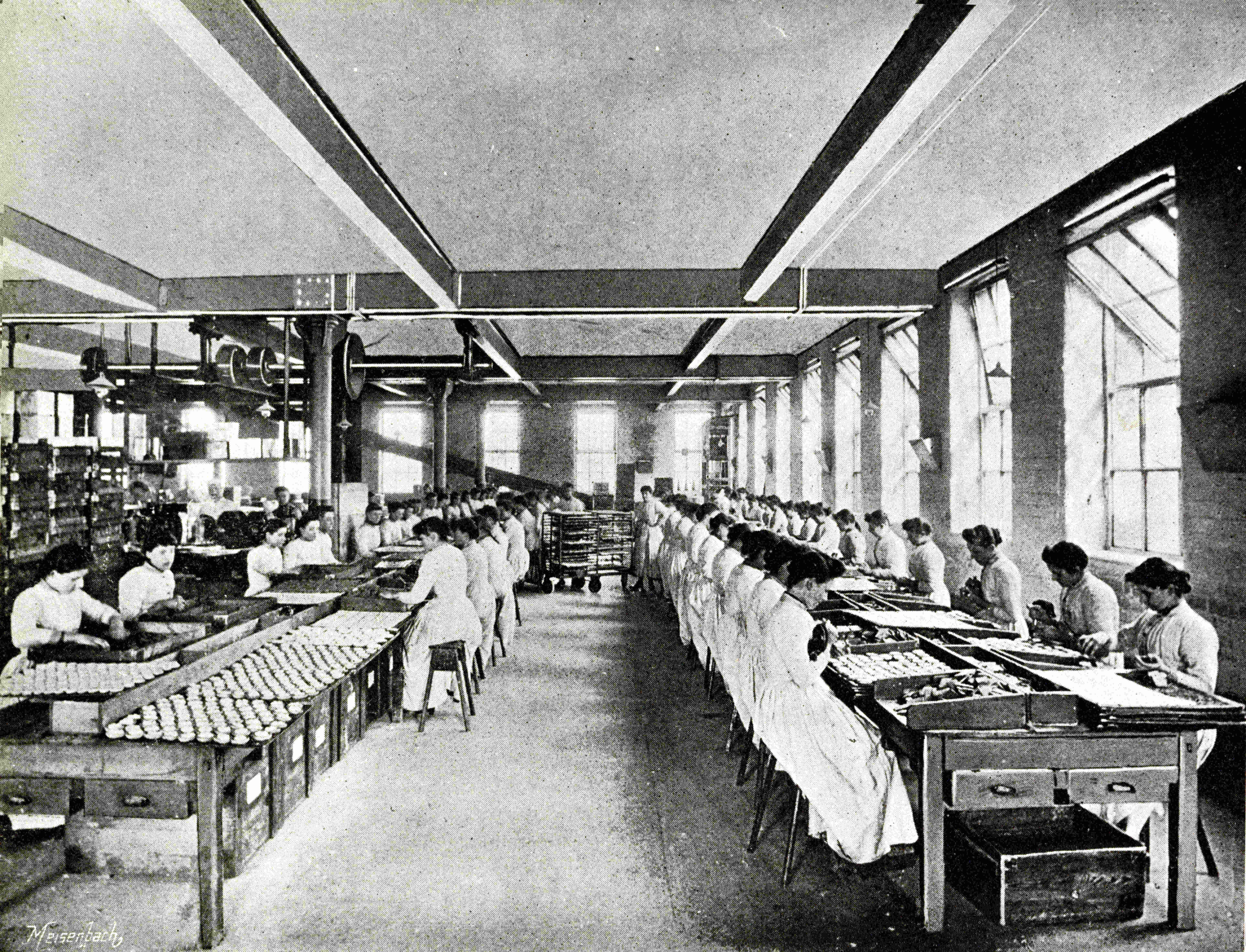 A Victorian photograph of a long production hall with rows of women in white aprons working at benches on either side, biscuits visible on the left-hand line