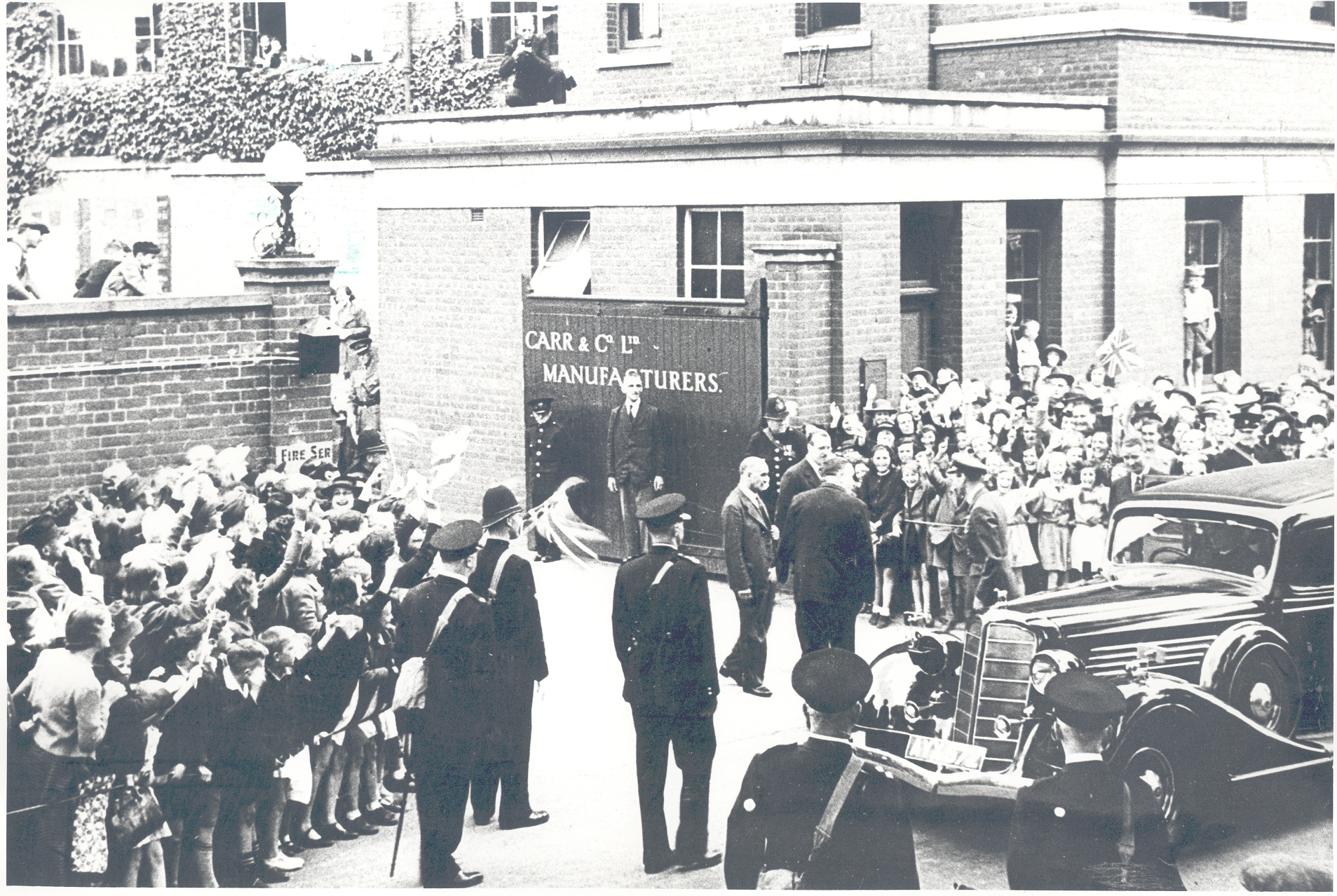 A black-and-white photograph of a royal car arriving at Carr and Co Ltd Manufacturers, with an eager crowd of children and adults lining the street