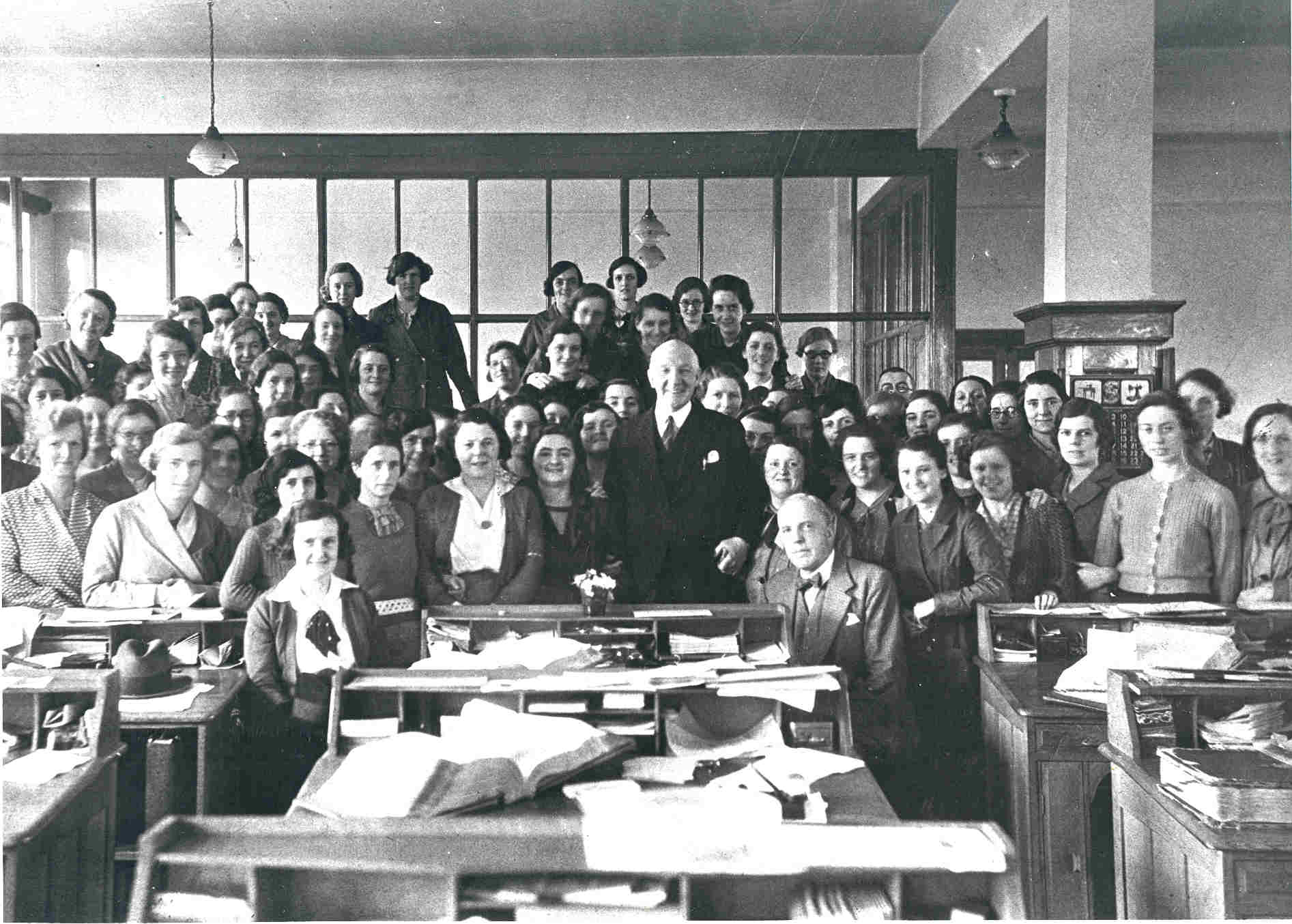 A 1930s photograph of predominantly female factory office staff gathered around desks, with a senior manager standing in the centre